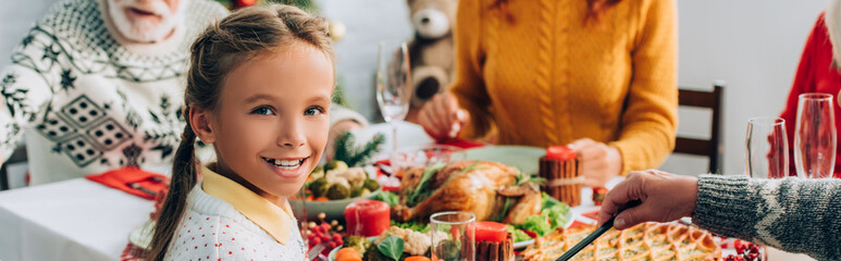 Panoramic crop of girl looking at camera, sitting at festive table near family