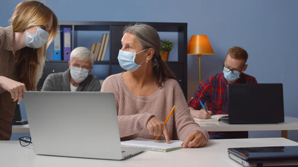 Teacher in mask helping mature student in class working at computer