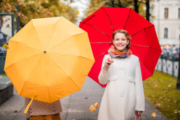 Girls with red and yellow umbrellas walking towards each other on autumn street of the city. Dry fallen leaves on the road.