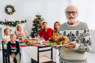 happy senior man in eyeglasses holding plate with tasty roasted turkey during festive dinner with family
