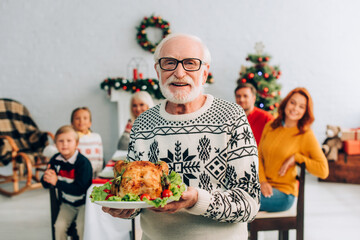 happy senior man in eyeglasses holding tasty roasted turkey during festive dinner with family