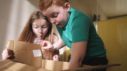 Happy little sister and brother opening cardboard box and pulling out gift from it while at room. Amazed small girl and boy showing joy and surprise on their faces. Cute children enjoying to present
