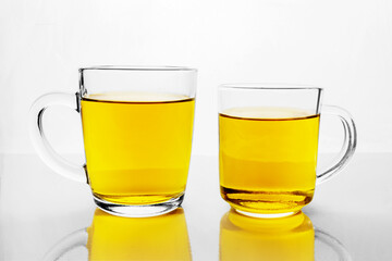 Two transparent glass cups with yellow liquid on white reflective surface. Mugs of tea or juice.