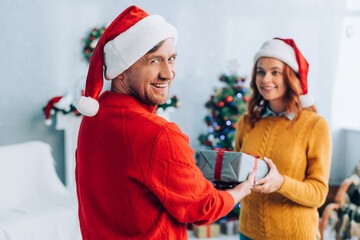 cheerful man looking at camera while presenting gift box to smiling wife