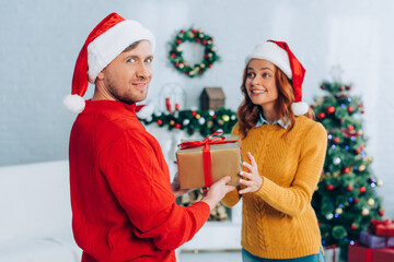 smiling man in santa hat looking at camera while presenting gift box to excited wife