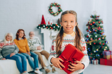 happy girl holding gift box and looking at camera while mother and grandparents sitting on background