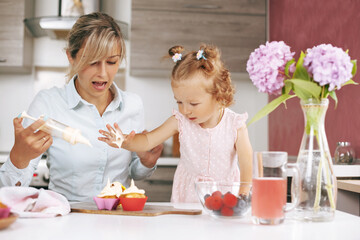 A beautiful young girl and her little daughter decorate freshly baked cupcakes with cream. On the table are cupcakes, berries, juice and a bouquet of hydrangeas. Family vacation concept