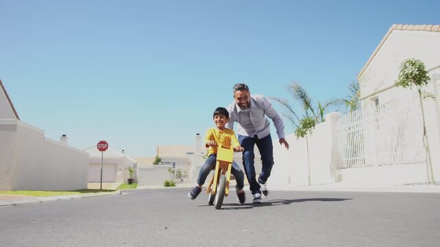Happy latin father helping smiling boy to ride wooden balance cycle on street. Happy middle eastern child and young dad riding bike in lane. Smiling daddy teaching son to ride a balance bicycle.