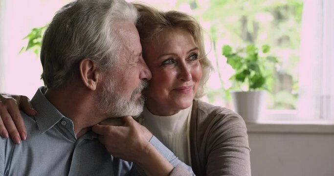 Close up happy elderly spouses sit together indoor hold hands day dreaming looking into distance. Loving husband kiss on cheek beloved 55s wife. Eternal love, welfare retired couple portrait concept