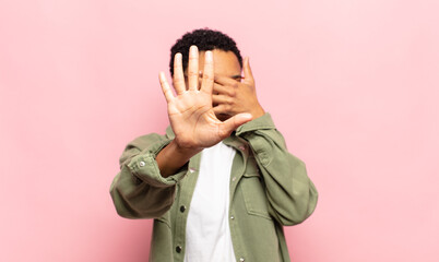 afro young black woman covering face with hand and putting other hand up front to stop camera,...