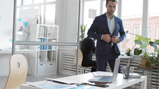 Medium long of middle-aged Caucasian male business professional entering office, unbuttoning blazer, sitting down on chair by desk and starting working on laptop, colleague in background 