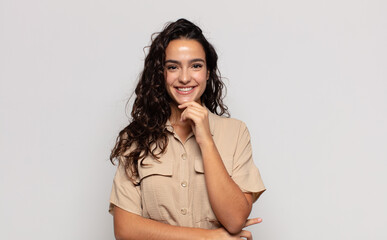 pretty young woman looking happy and smiling with hand on chin, wondering or asking a question,...