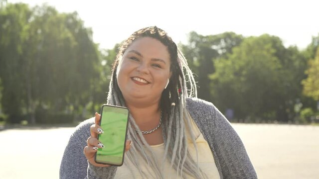 Young pretty fat woman with dreads showing green screen phone to camera and dancing. Beautiful stout girl with chromakey on her smartphone advertising mobile app. Commercial, copy space concept.