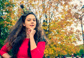 Girl brunette in a fairytale image in an autumn park. A girl in a red dress and a black vest against a yellow leaves licks her lips threatening to bite. Insidious cunning witch looking to the camera