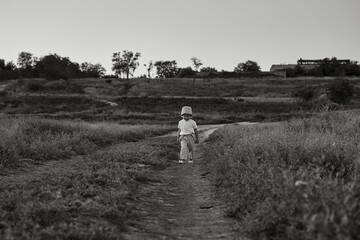 Two-year-old child in a hat walks along a country road, summer. Knowledge of the world. Black and white photo