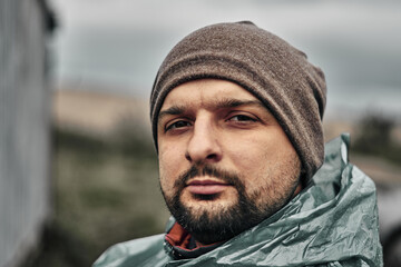 A man with a beard in a green raincoat and hat, face close-up. Overcast weather.