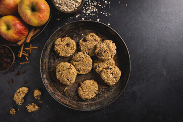 Freshly baked apple, oatmeal, raisin, cinnamon and mixed nuts cookies with ingredients on a dark concrete surface. Room for text.