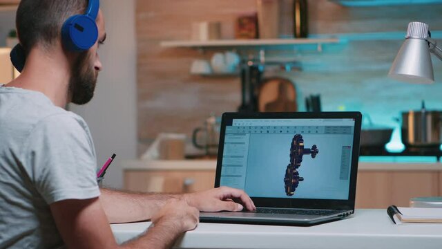 Man architect with wireless headset using laptop while working at home at night sitting in the kitchen. Industrial employee studying prototype idea on personal computer showing cad software on display