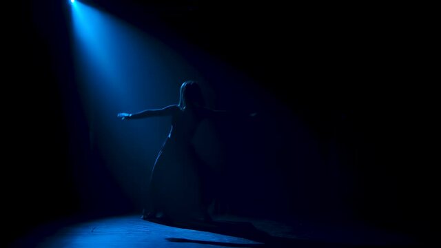 Silhouette of a graceful black robed dancer doing a series of dance steps and acrobatic elements during a contemporary contemp performance in a beam of blue light. Slow motion