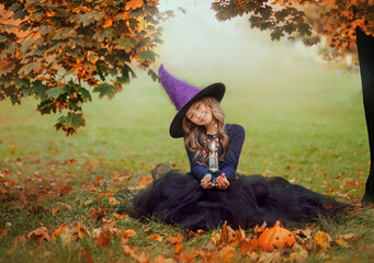A little beautiful girl in a witch costume sits on the green grass and holds a burning candle,...