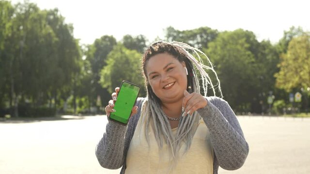 Beautiful stout girl with chromakey on her smartphone advertising mobile app. Young pretty fat woman with dreads showing green screen phone to camera and dancing. Commercial, copy space concept.