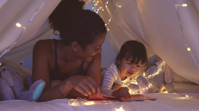 African american mother lying on bed in illuminated tent and playing with cute girl. Close up of mom telling a fairy tale in kid tent. Mother reading a story to little daughter before going to bed.