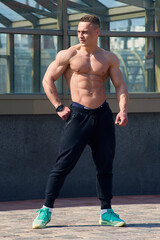 A muscular shirtless man is posing full length near the structure at noon. A shredded bodybuilder...