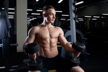 Fototapeta na wymiar A muscular shirtless man is holding dumbbells on his knees in a gym. A bodybuilder is doing a workout for getting ready for the bodybuilding competition. A guy is on a pre-competitive diet.