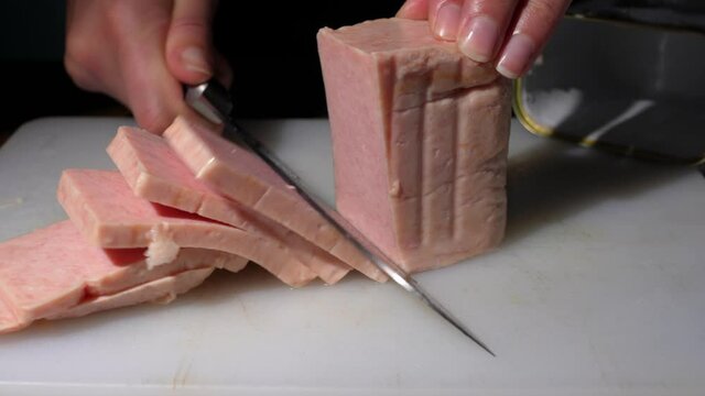 This close up video shows anonymous hands cutting and slicing a block of canned ham on a cutting board with a kitchen knife. 