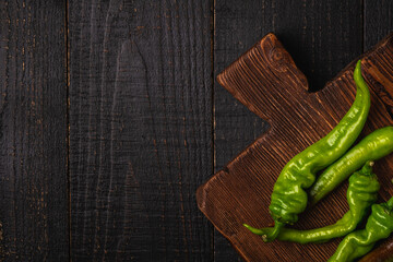 Hot spicy green chili peppers on teak wooden cutting board, dark wood backdrop background, top view copy space