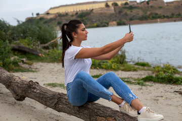 Fototapeta na wymiar A young woman is sitting on the riverbank and taking a selfie 