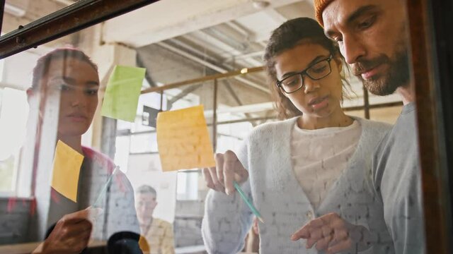 Three young business people discussing and planning strategy in front of glass board and stickers, slow motion