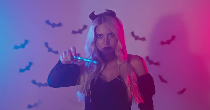 Mysterious woman in Halloween costume and horns gesturing with magic wand, smoky studio with neon lights