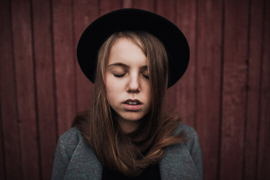 Serious young blonde teenager girl with long hair and freckles on face in fashionable black felt hat.eyes closed. the mouth is open.hair is twisted around the neck.Red wooden shabby striped background