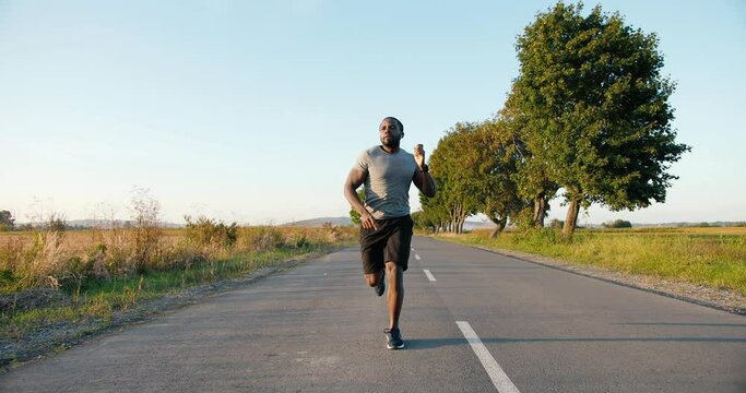 African American handsome strong male jogger in headphones running on road in countryside on summer day. Attractive sporty man jogging outdoor. Sport concept. Sportsman runner. Morning nature.