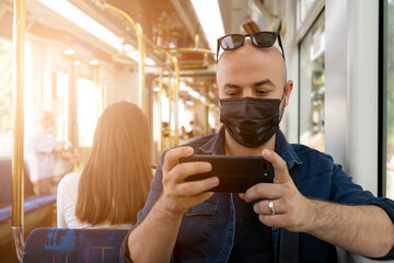 A young man in a black mask looking at his smartphone while traveling on the subway.