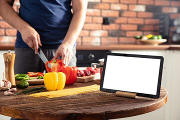 Tablet on the kitchen table and man preparing delicious and healthy food on background. Tablet screen with empty space.