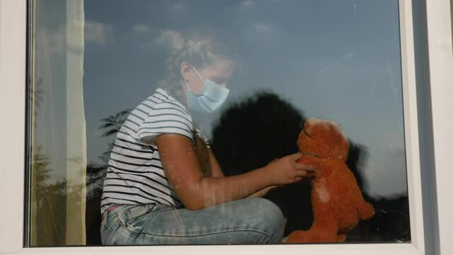 bored kid in medical mask in home quarantine coronavirus sitting by the window. child with covid 19 a toy teddy bear in protective mask looking out the window. coronavirus epidemic prevention concept