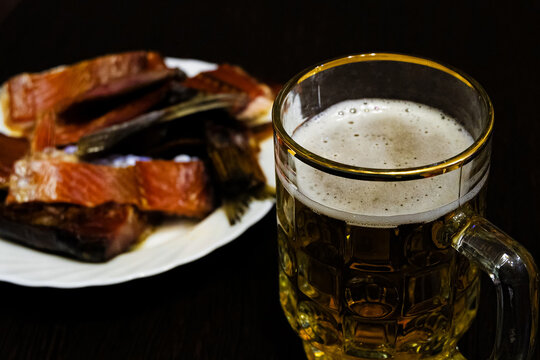 Glass half-liter mug filled with beer on the blurred background of a snack of dried smoked fish on the table. Selective focus on glass with beer.