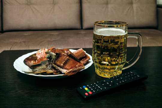 Glass half-liter mug filled with beer, dried smoked fish and TV remote control on the table. Glass with beer and snack against the backdrop of the sofa.