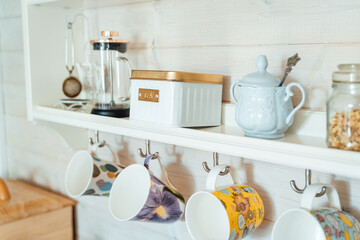 Obraz na płótnie Canvas Kitchen wooden shelf with tea leaves in gold box and accessories, blue sugar bowl with spoon, strainer, press. Many colorful cups, mugs are hanging from hooks. Cozy interior in a country house