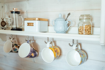 Fototapeta na wymiar Kitchen wooden shelf with tea leaves in gold box and accessories, blue sugar bowl with spoon, strainer, press. Many colorful cups, mugs are hanging from hooks. Cozy interior in a country house