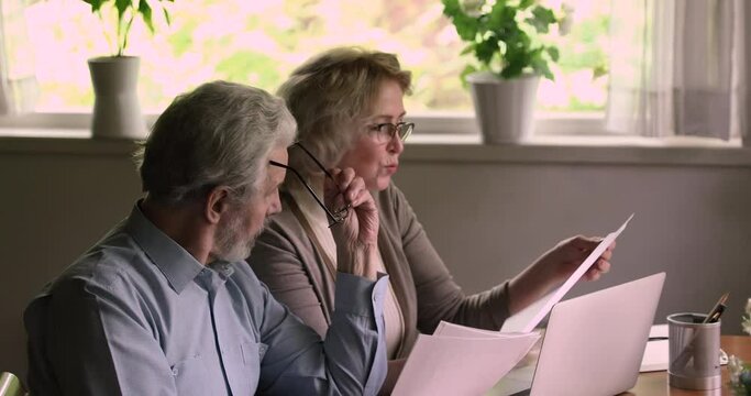 Sixty years old wife and husband sit at table holding sheets reading bank loan agreement, manage family budget, check utility bills, discuss medical insurance coverage conditions terms feels satisfied