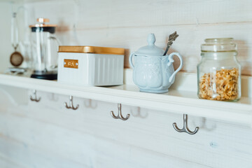 Kitchen wooden shelf with tea leaves in gold box and accessories, blue sugar bowl with spoon, strainer, press. Cup, mug hooks. Cozy interior in a country house
