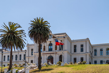Obraz premium Valparaiso, Chile - December 8, 2008: White historic building of Navy Headquarters on green lawn hill under blue sky with couple of palm trees and a few machine guns. Flag adds color.