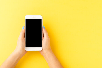 Woman’s hands using mobile phone with empty screen on yellow background. Top view