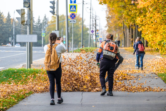A utility worker uses a blower to remove fallen leaves in an alley in a park. Yellow leaves are flying in the air. The girl behind takes a picture of the worker using a smartphone. Seasonal work.
