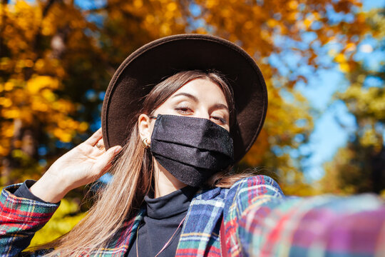 Portrait of woman wearing mask in fall park during coronavirus covid-19 pandemic and taking selfie. Stylish autumn