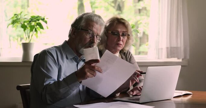 Aged couple sit at table use laptop analyzing domestic bills arguing about unreasonable expenses money overspend. Old citizens with small pension, bankruptcy, lack of money financial problems concept