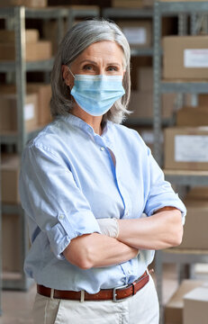 Older Business Woman Retail Seller, Entrepreneur, Small Business Owner Wearing Face Mask And Gloves For Covid 19 Protection Looking At Camera Standing In Delivery Storage Warehouse, Vertical Portrait.